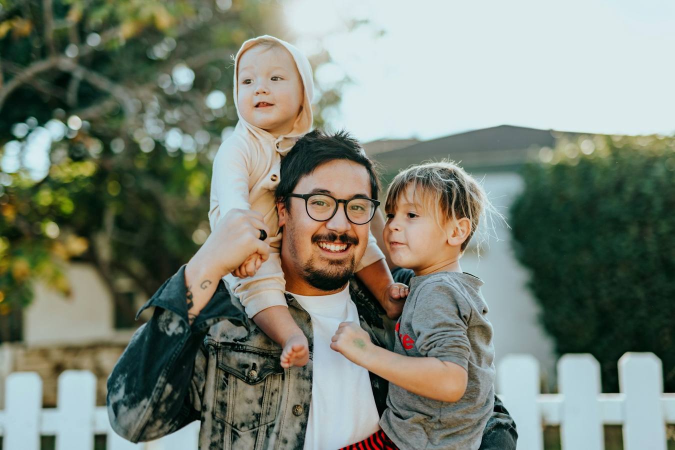 Parent smiling with two children beside a white fence and family home